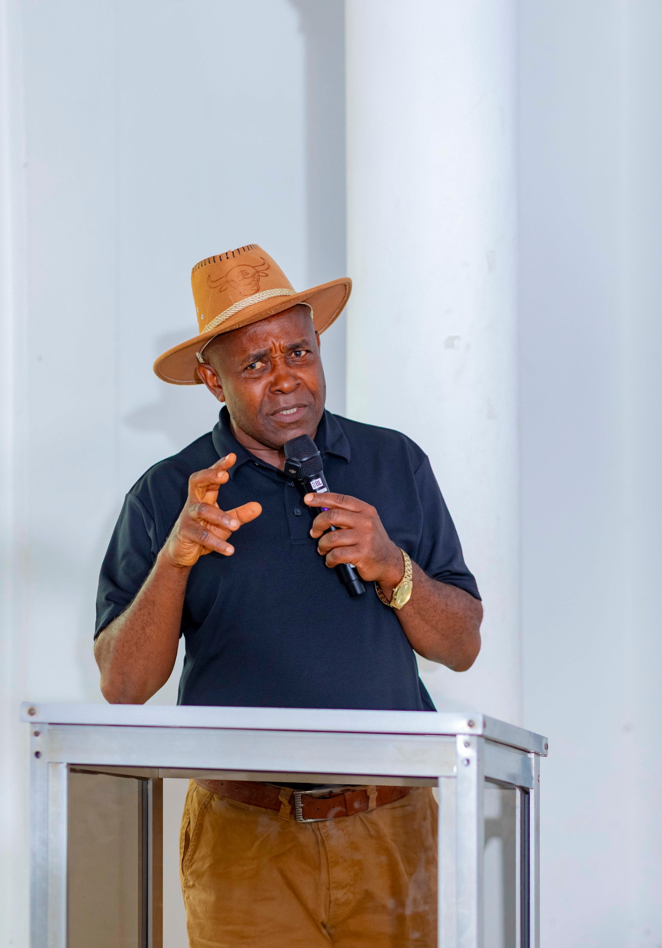 Conference facilitator in a hat addressing participants from the podium during the GRA Inaugural Colloquium