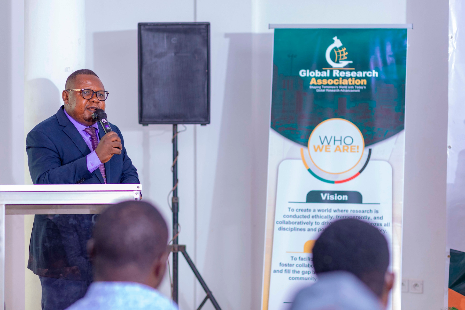 Conference facilitator speaking at the podium with the GRA mission banner in view during the GRA Inaugural Colloquium