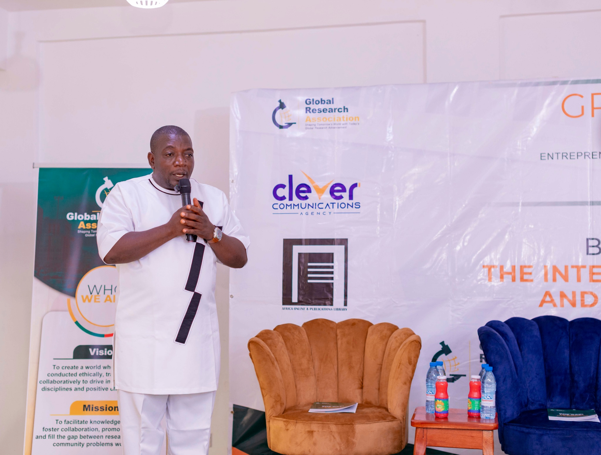 Conference facilitator in white attire speaking on stage during the GRA Inaugural Colloquium