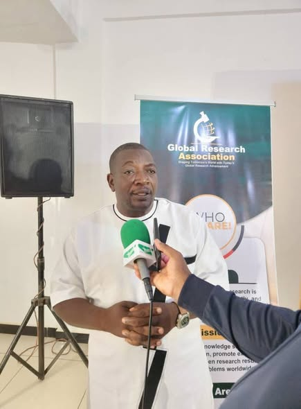 GRA representative speaking into a press microphone in front of the Global Research Association banner